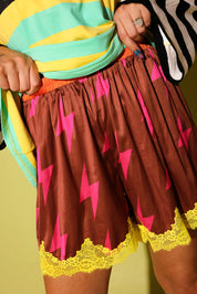 Close up of a a model wearing maroon lace shorts with pink lightening bolts and yellow lace trim holding waistband, and colorful striped shirt, against a pale yellow background
