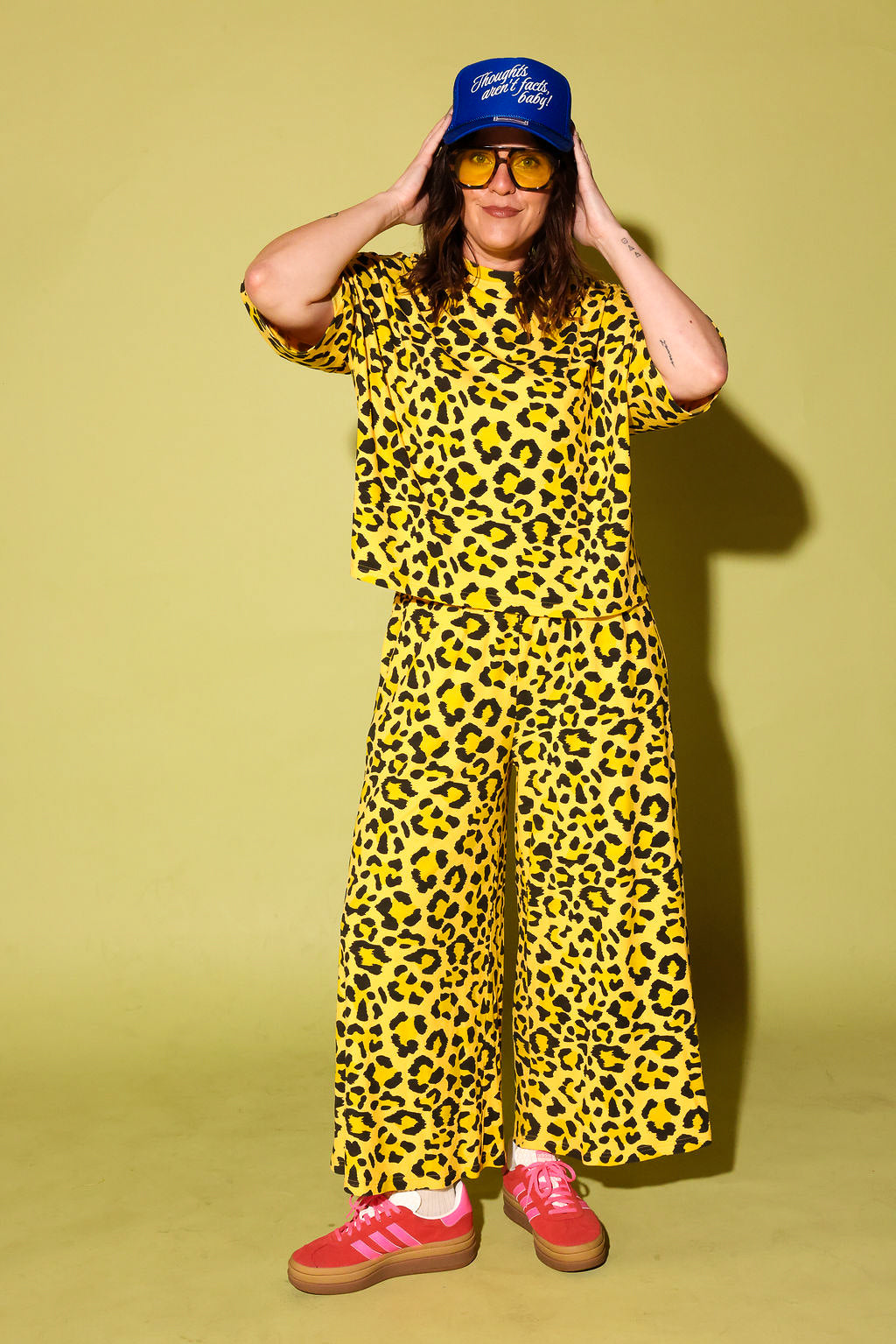 Model wearing a black and yellow leopard set black high heels and a blue trucker hat with text on it, standing against a pale yellow background