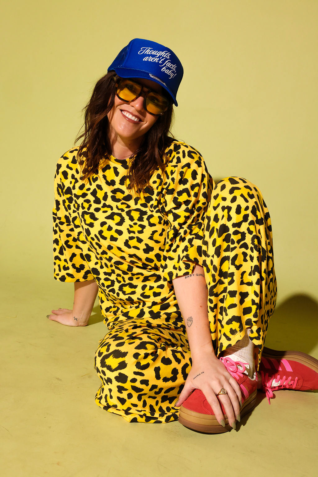 Model wearing a black and yellow leopard set black high heels and a blue trucker hat with text on it, sitting against a pale yellow background