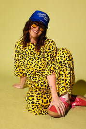 Model wearing a black and yellow leopard set black high heels and a blue trucker hat with text on it, sitting against a pale yellow background