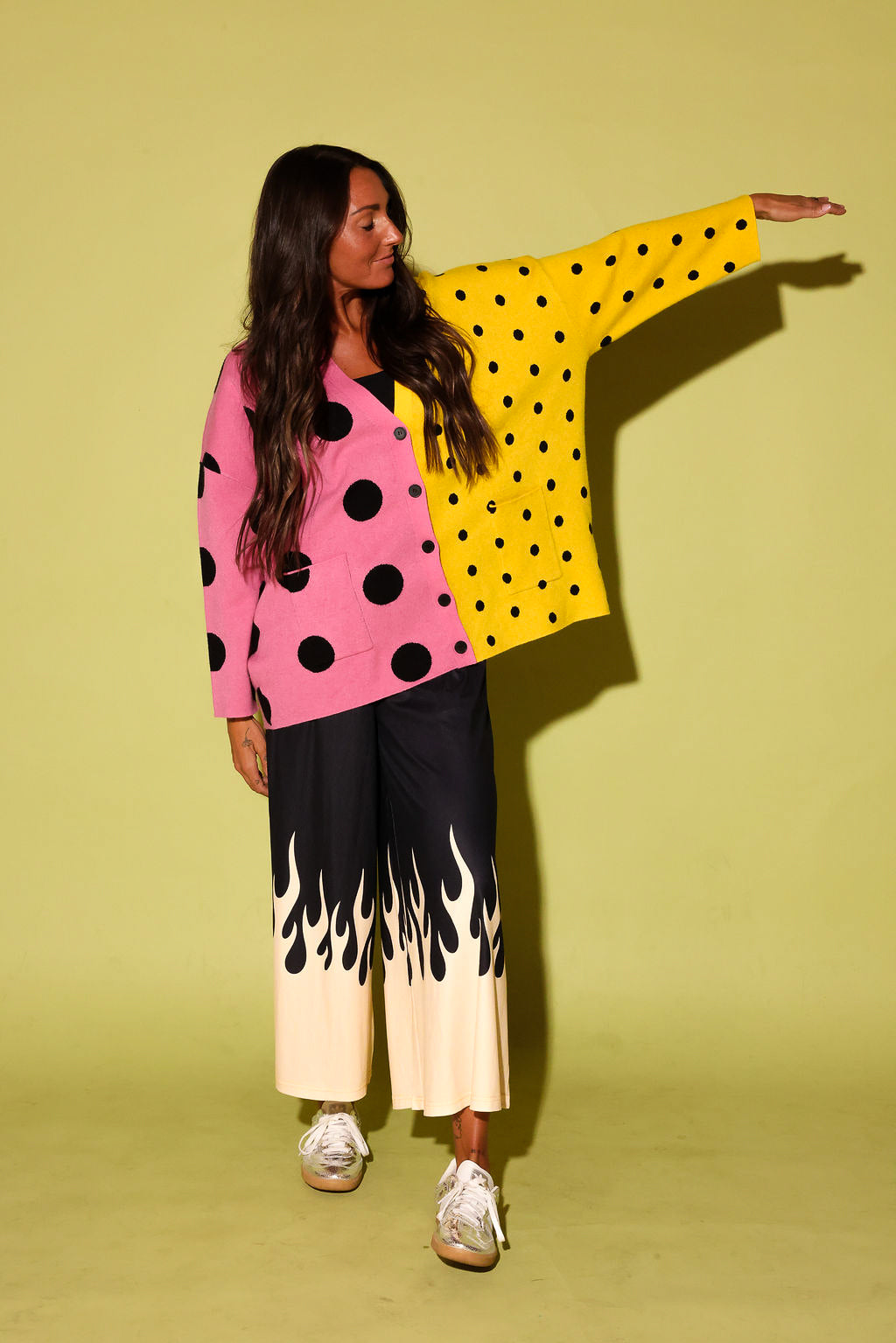 A model posing in a vibrant, split-color polka dot cardigan with pink and yellow halves, paired with black wide-leg pants featuring a cream flame pattern at the hem, set against a solid pale yellow-green studio backdrop.