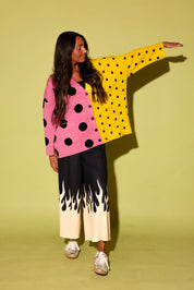 A model posing in a vibrant, split-color polka dot cardigan with pink and yellow halves, paired with black wide-leg pants featuring a cream flame pattern at the hem, set against a solid pale yellow-green studio backdrop.