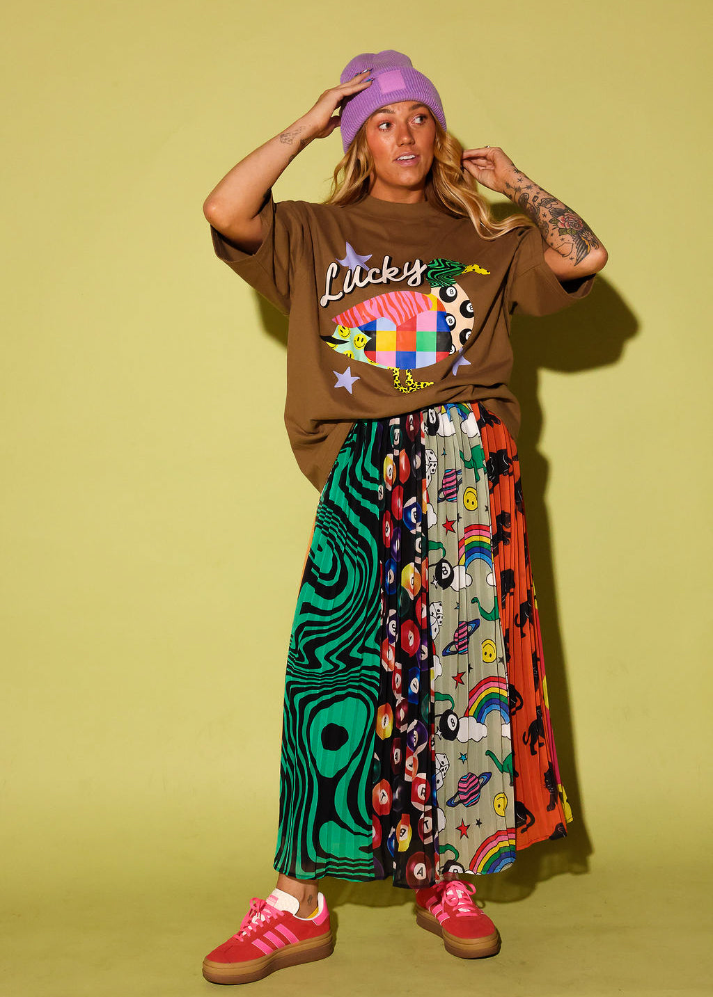A model stands against a solid pale yellow-green studio backdrop wearing a brown graphic t-shirt with 'Lucky' written on it, a long, wide-leg pleated skirt with multiple colorful patterned panels, a purple beanie, and pink sneakers.