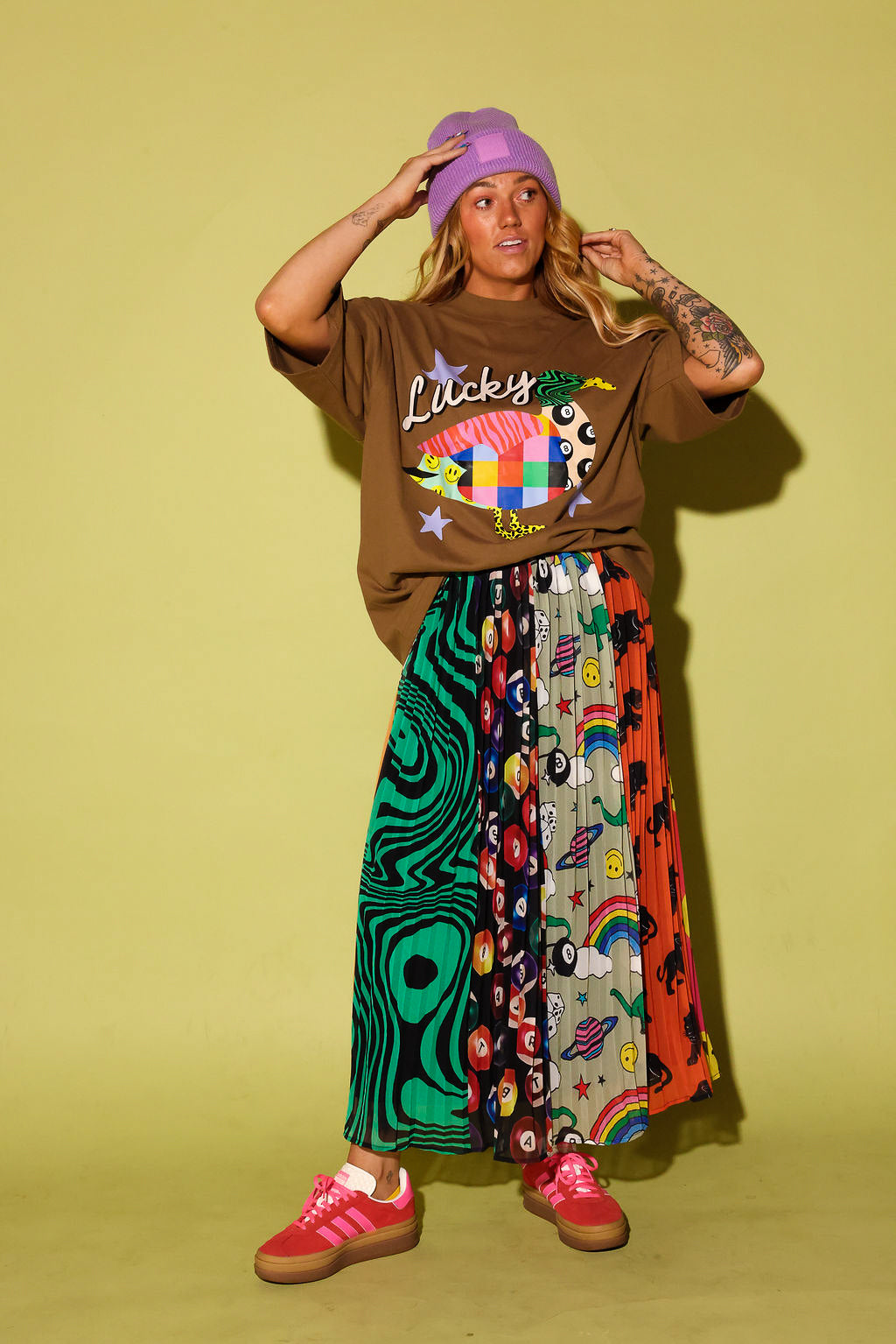 A model stands against a solid pale yellow-green studio backdrop wearing a brown graphic t-shirt with 'Lucky' written on it, a long, wide-leg pleated skirt with multiple colorful patterned panels, a purple beanie, and pink sneakers.