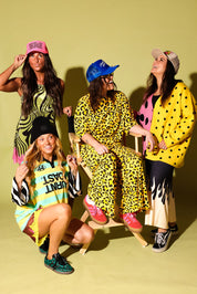 Four women posing together in vibrant, colorful outfits with bold patterns like leopard print, polka dots, and stripes, set against a solid pale yellow-green studio background.