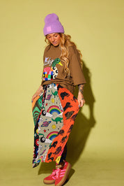 A model wearing a brown graphic t-shirt, a purple beanie, and a long, colorful skirt with dinosaur and rainbow prints, paired with pink sneakers, set against a solid yellow-green studio background.