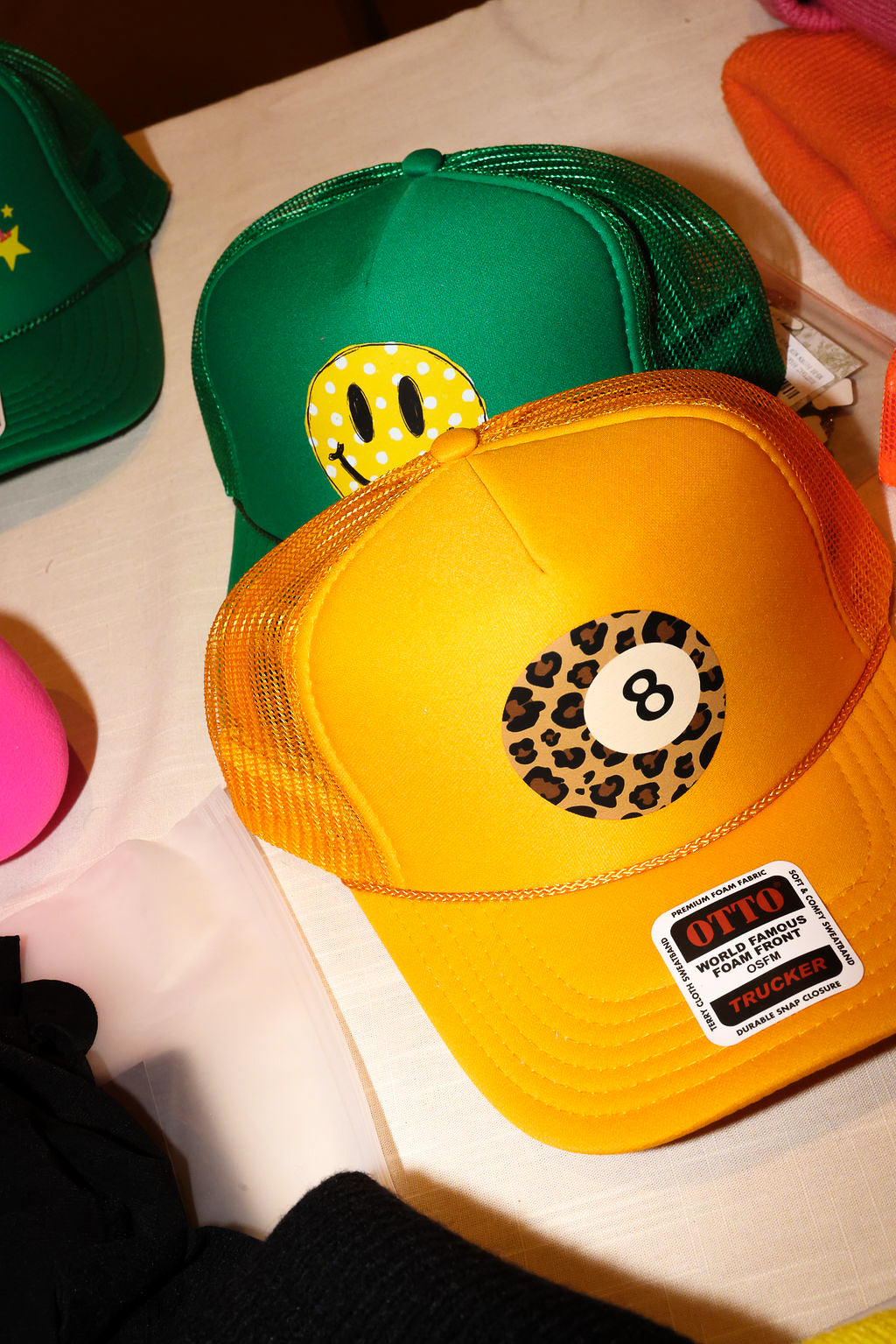 A yellow and orange trucker hat with a leopard-print 8-ball graphic on the front, layered over a green trucker hat with a yellow smiley face patch. Both hats feature a visible 'OTC' brand sticker on the brim, set against a white tabletop background.