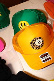A yellow and orange trucker hat with a leopard-print 8-ball graphic on the front, layered over a green trucker hat with a yellow smiley face patch. Both hats feature a visible 'OTC' brand sticker on the brim, set against a white tabletop background.
