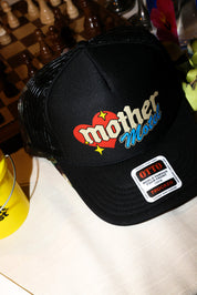 A black trucker-style baseball cap featuring a retro graphic on the front with a red heart, yellow stars, and the words 'mother Motel' in beige and blue lettering. A visible patch on the brim reads 'OTTO World Famous Trucker Hats.