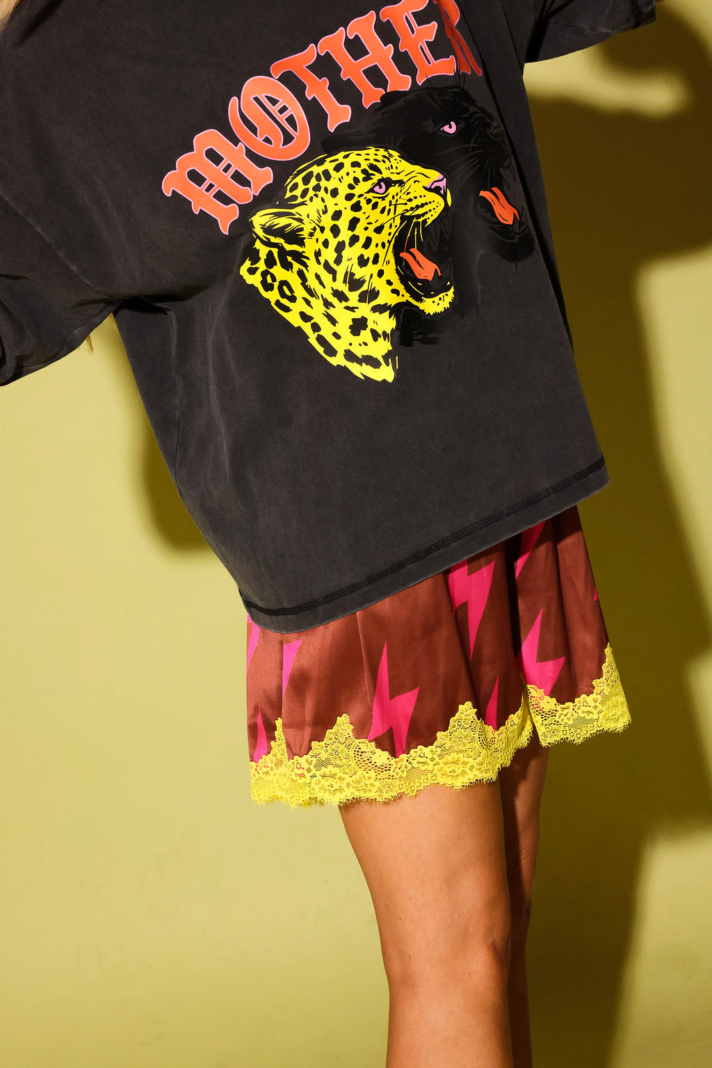 Close up of a grey graphic tee with the word 'MOTHER' and 2 panthers and red lace shorts with pink lightening bolts and yellow lace on a pale yellow background