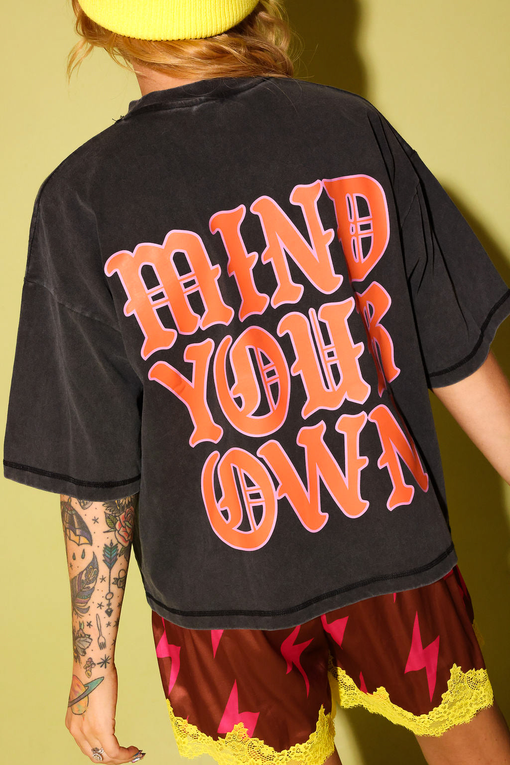 The back of a model wearing a grey shirt with the text 'MIND YOUR OWN' in pink and orange and maroon lace shorts, and a yellow beanie, on a pale yellow background