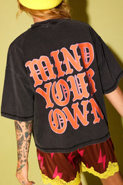 The back of a model wearing a grey shirt with the text 'MIND YOUR OWN' in pink and orange and maroon lace shorts, and a yellow beanie, on a pale yellow background