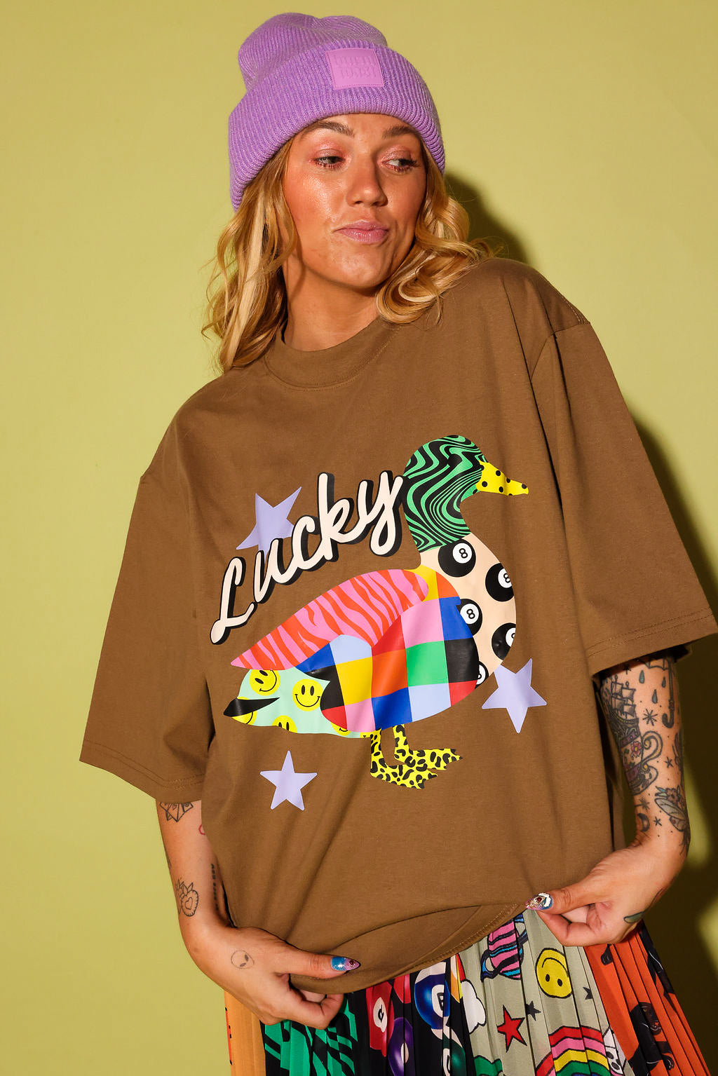 A woman wearing an oversized brown graphic t-shirt with a colorful duck design and the word 'Lucky' in script, paired with a purple beanie and a patterned skirt, set against a solid olive green background.