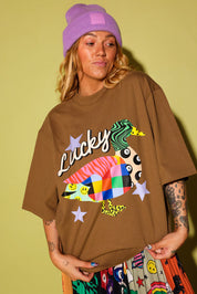 A woman wearing an oversized brown graphic t-shirt with a colorful duck design and the word 'Lucky' in script, paired with a purple beanie and a patterned skirt, set against a solid olive green background.