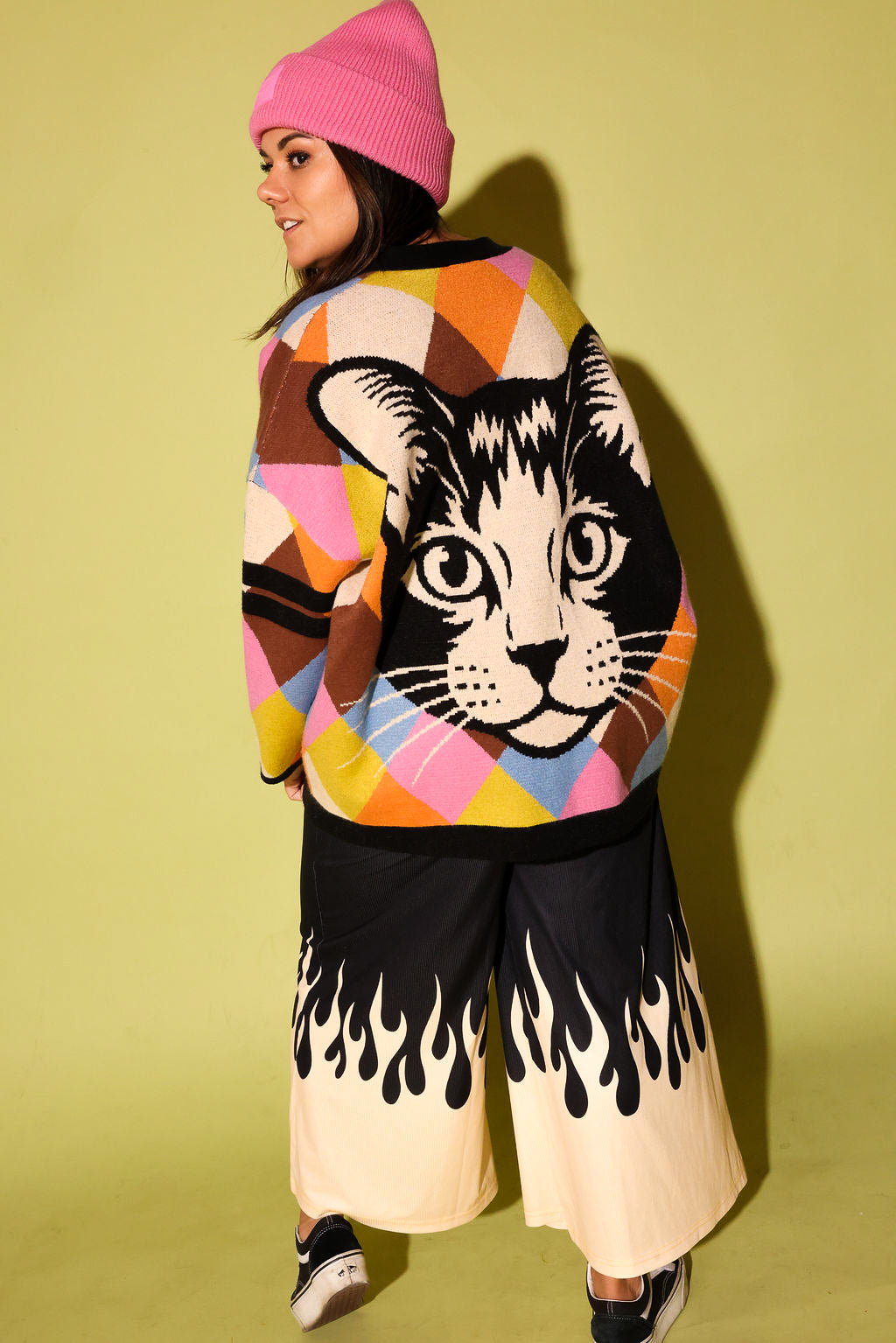 A model wearing a multicolored diamond pattern cardigan with a cat graphic on the back with a black pants white flames in creme, pink beanie beanie, and black  sneakers, on a pale green background