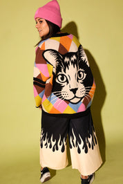 A model wearing a multicolored diamond pattern cardigan with a cat graphic on the back with a black pants white flames in creme, pink beanie beanie, and black  sneakers, on a pale green background