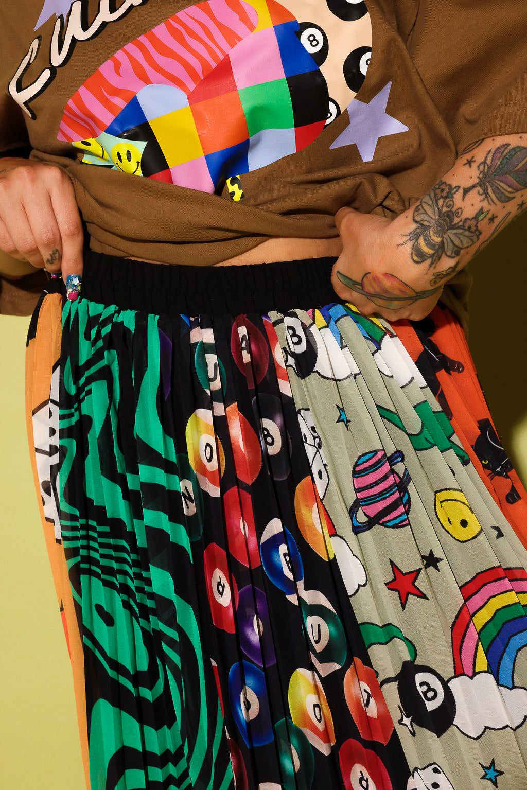 A close-up of a person wearing a brown graphic sweatshirt and a pleated skirt with multiple colorful panels featuring billiards, planets, stars, and abstract patterns.