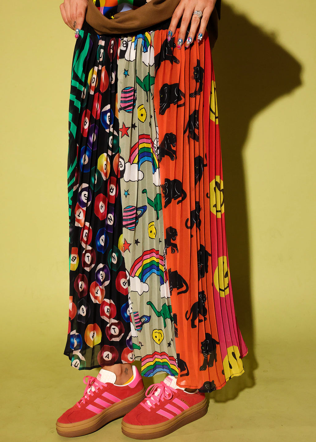A person wearing a long pleated maxi skirt with multiple colorful printed panels, including black-and-white floral, rainbow, dino, and smiley-face motifs, paired with pink sneakers against a yellow-green studio background.
