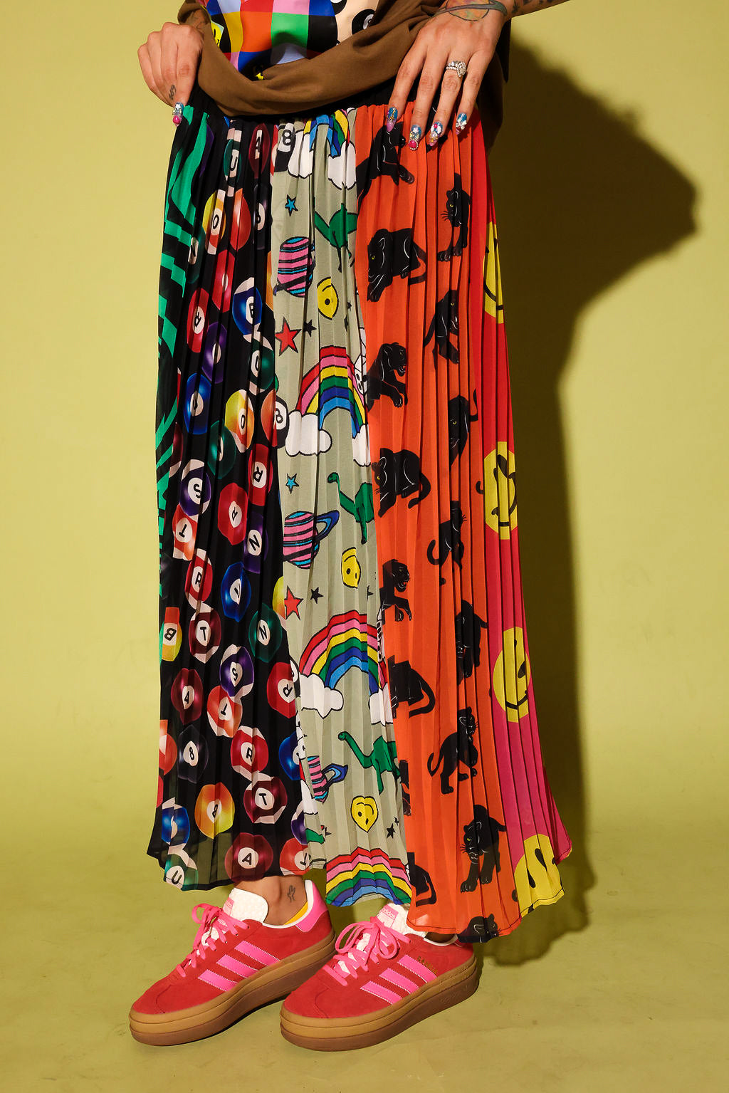 A person wearing a long pleated maxi skirt with multiple colorful printed panels, including black-and-white floral, rainbow, dino, and smiley-face motifs, paired with pink sneakers against a yellow-green studio background.