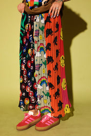 A person wearing a long pleated maxi skirt with multiple colorful printed panels, including black-and-white floral, rainbow, dino, and smiley-face motifs, paired with pink sneakers against a yellow-green studio background.