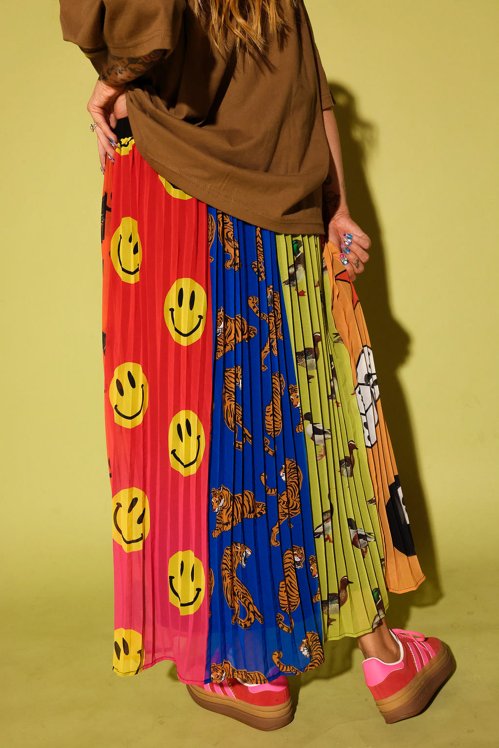 A person wearing a long pleated skirt with multiple vertical color-blocked panels featuring smiley faces, tigers, and cartoon graphics, paired with a brown top and pink platform sneakers against a yellow-green studio background.