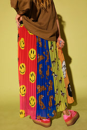 A person wearing a long pleated skirt with multiple vertical color-blocked panels featuring smiley faces, tigers, and cartoon graphics, paired with a brown top and pink platform sneakers against a yellow-green studio background.