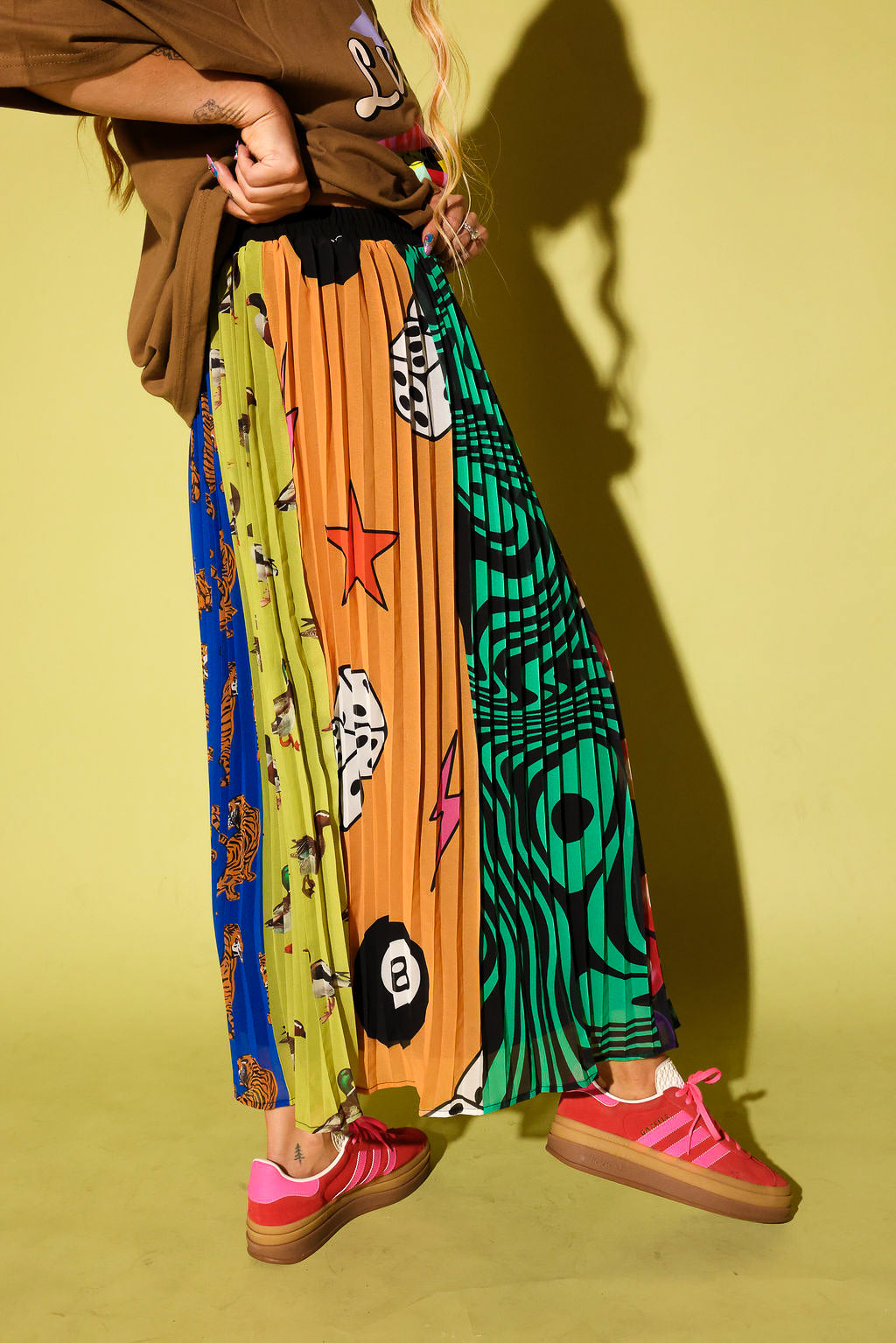 A person wearing a brown graphic t-shirt and a highly colorful pleated skirt with various patterns, paired with pink sneakers, standing against a yellow-green background with a strong shadow.