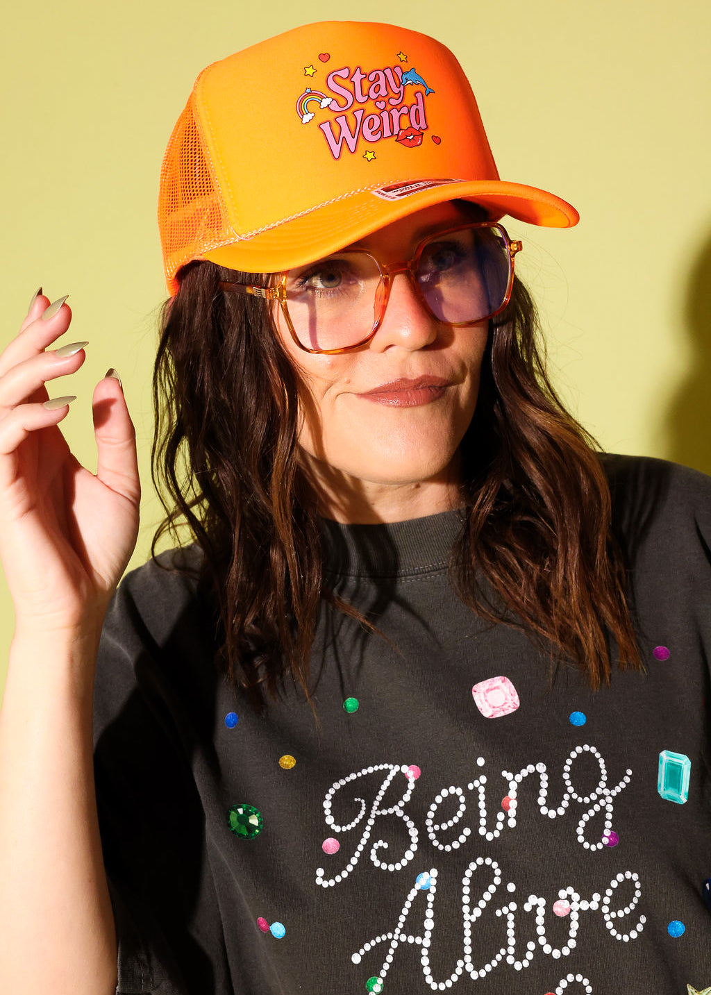Model wear a bright orange trucker hat with Stay Weird text in pink  and red and gray graphic tee on a pale yellow background