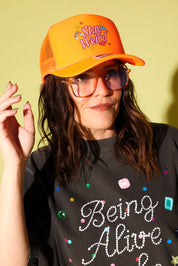 Model wear a bright orange trucker hat with Stay Weird text in pink  and red and gray graphic tee on a pale yellow background