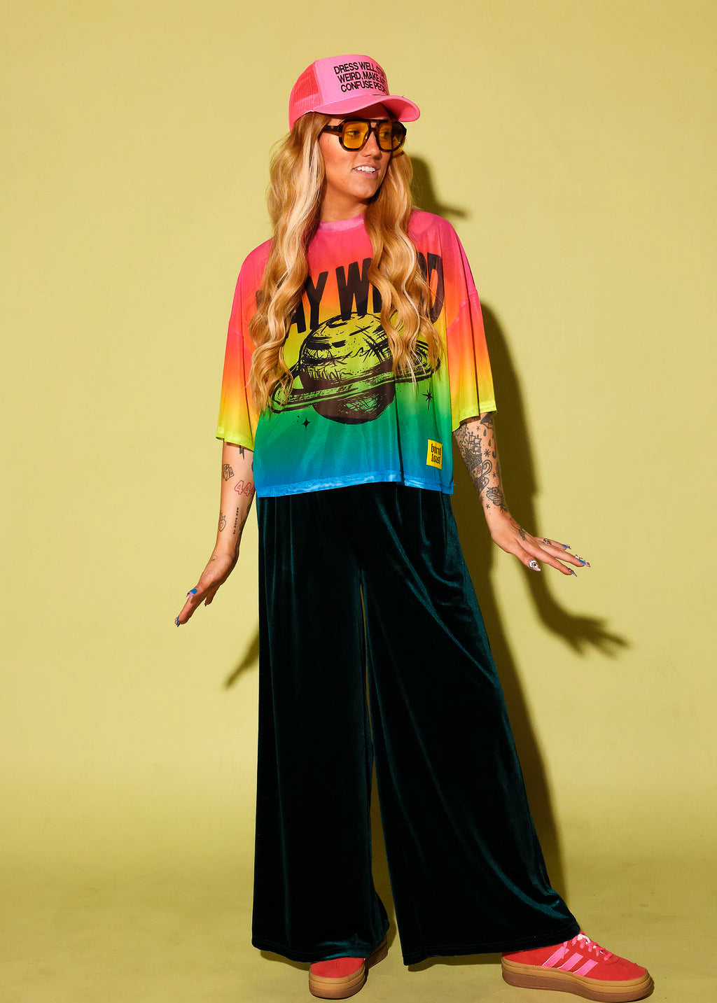 A model wearing a rainbow gradient mesh top with the text 'STAY WEIRD' and a planet in black and green velvet pants, pink sneakers and neon pink trucker hat, against a pale yellow background