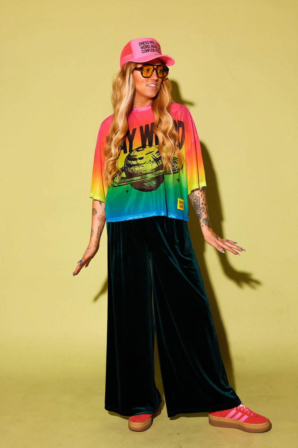 A model wearing a rainbow gradient mesh top with the text 'STAY WEIRD' and a planet in black and green velvet pants, pink sneakers and neon pink trucker hat, against a pale yellow background
