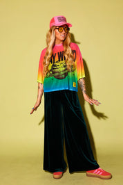 A model wearing a rainbow gradient mesh top with the text 'STAY WEIRD' and a planet in black and green velvet pants, pink sneakers and neon pink trucker hat, against a pale yellow background