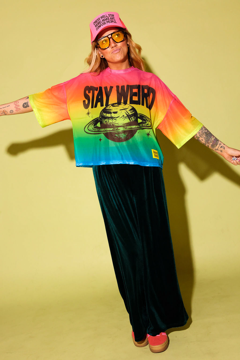A model wearing a rainbow gradient mesh top with the text 'STAY WEIRD' and a planet in black and green velvet pants, pink sneakers and neon pink trucker hat, against a pale yellow background