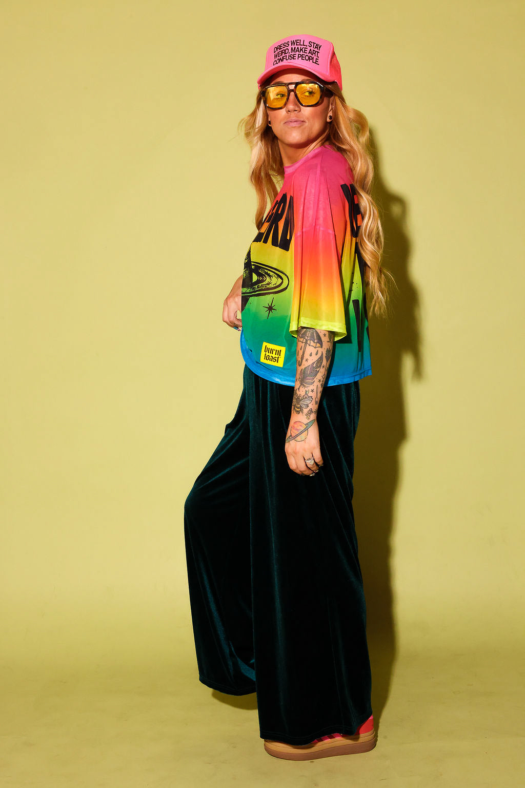Side view of a model wearing a rainbow gradient mesh top with the text 'STAY WEIRD' and a planet in black and green velvet pants, pink sneakers and neon pink trucker hat, against a pale yellow background