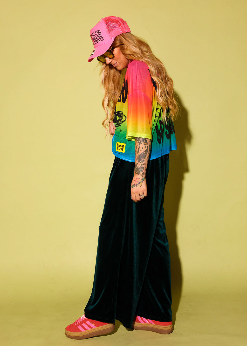 Side view of a model wearing a rainbow gradient mesh top with the text 'STAY WEIRD' and a planet in black and green velvet pants, pink sneakers and neon pink trucker hat, against a pale yellow background