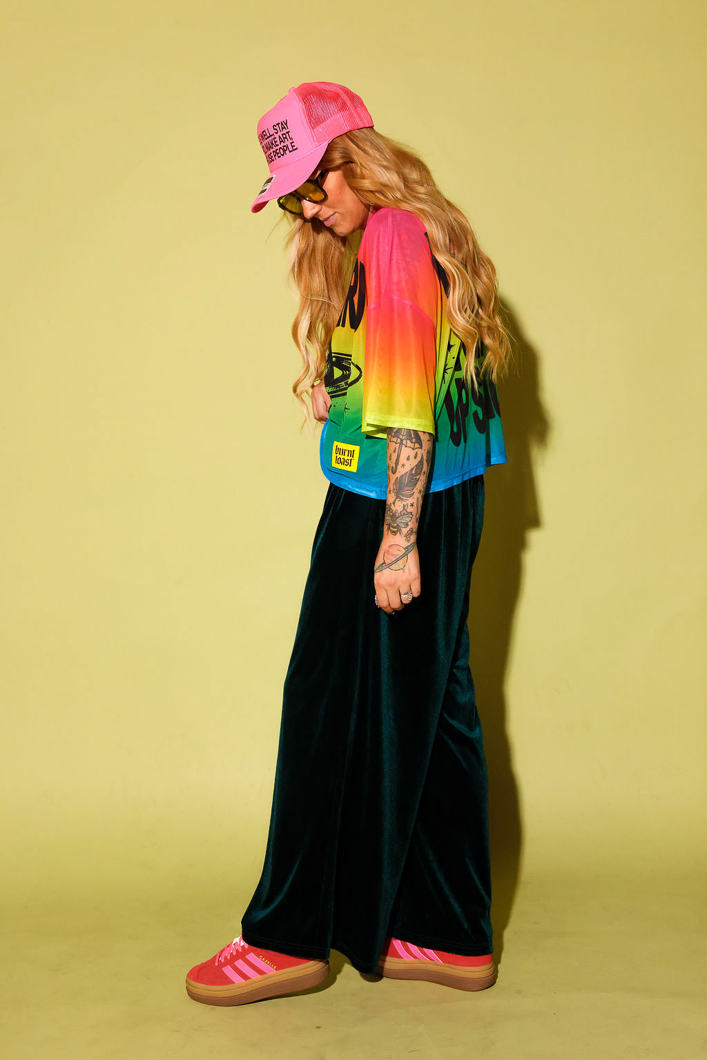 Side view of a model wearing a rainbow gradient mesh top with the text 'STAY WEIRD' and a planet in black and green velvet pants, pink sneakers and neon pink trucker hat, against a pale yellow background
