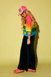 Side view of a model wearing a rainbow gradient mesh top with the text 'STAY WEIRD' and a planet in black and green velvet pants, pink sneakers and neon pink trucker hat, against a pale yellow background