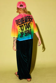 The back view of a model wearing a rainbow gradient mesh top with the text 'REMEMBER TO TAKE UP SPACE' in black and green velvet pants, pink sneakers and neon pink trucker hat, against a pale yellow background
