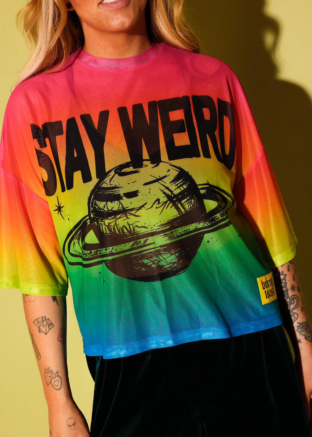 Close up of a model wearing a rainbow gradient mesh top with the text 'STAY WEIRD' and a planet in black and green velvet pants against a pale yellow background