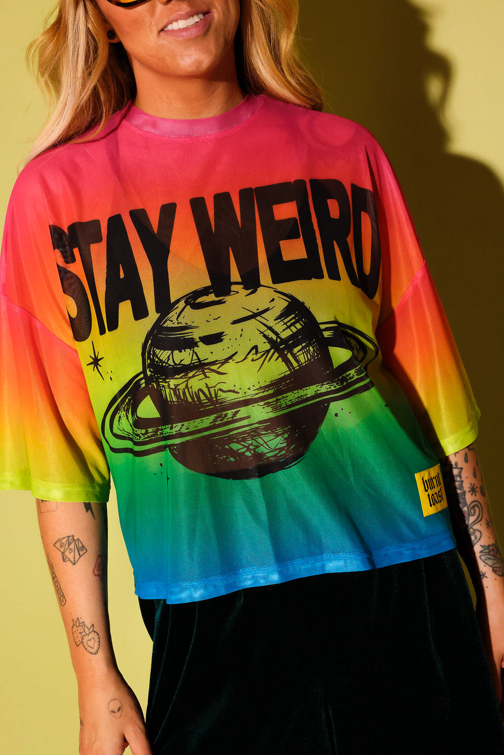 Close up of a model wearing a rainbow gradient mesh top with the text 'STAY WEIRD' and a planet in black and green velvet pants against a pale yellow background