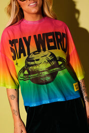 Close up of a model wearing a rainbow gradient mesh top with the text 'STAY WEIRD' and a planet in black and green velvet pants against a pale yellow background