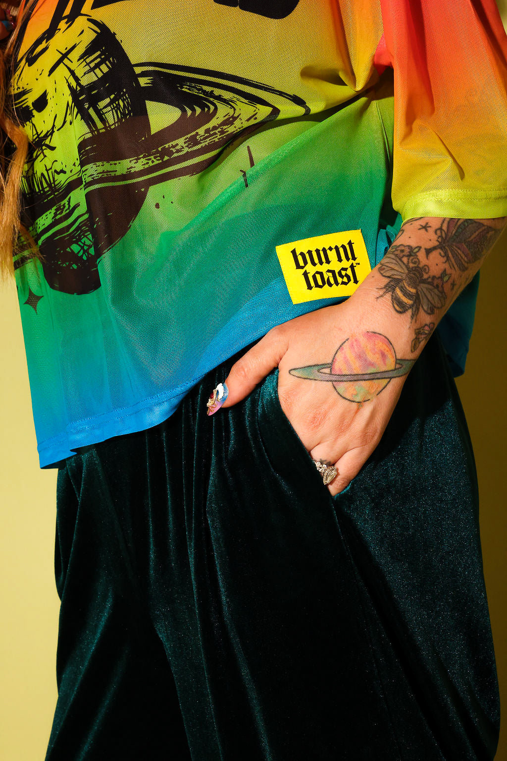 Close up side view of a model wearing a rainbow gradient mesh top with a planet in black and a hand in a pocket of green velvet pants, against a pale yellow background