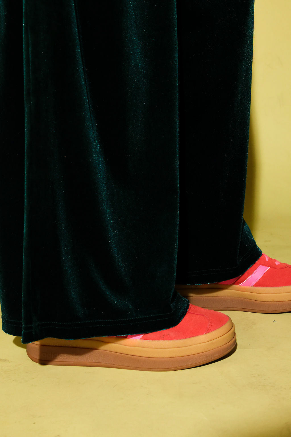 Close up view of the hem of green velvet pants and pink sneakers, against a pale yellow background