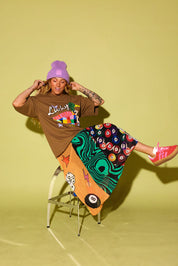 A model wearing a brown graphic t-shirt, a purple beanie, and a colorful patterned skirt sits on a metal stool against a solid yellow-green studio background.