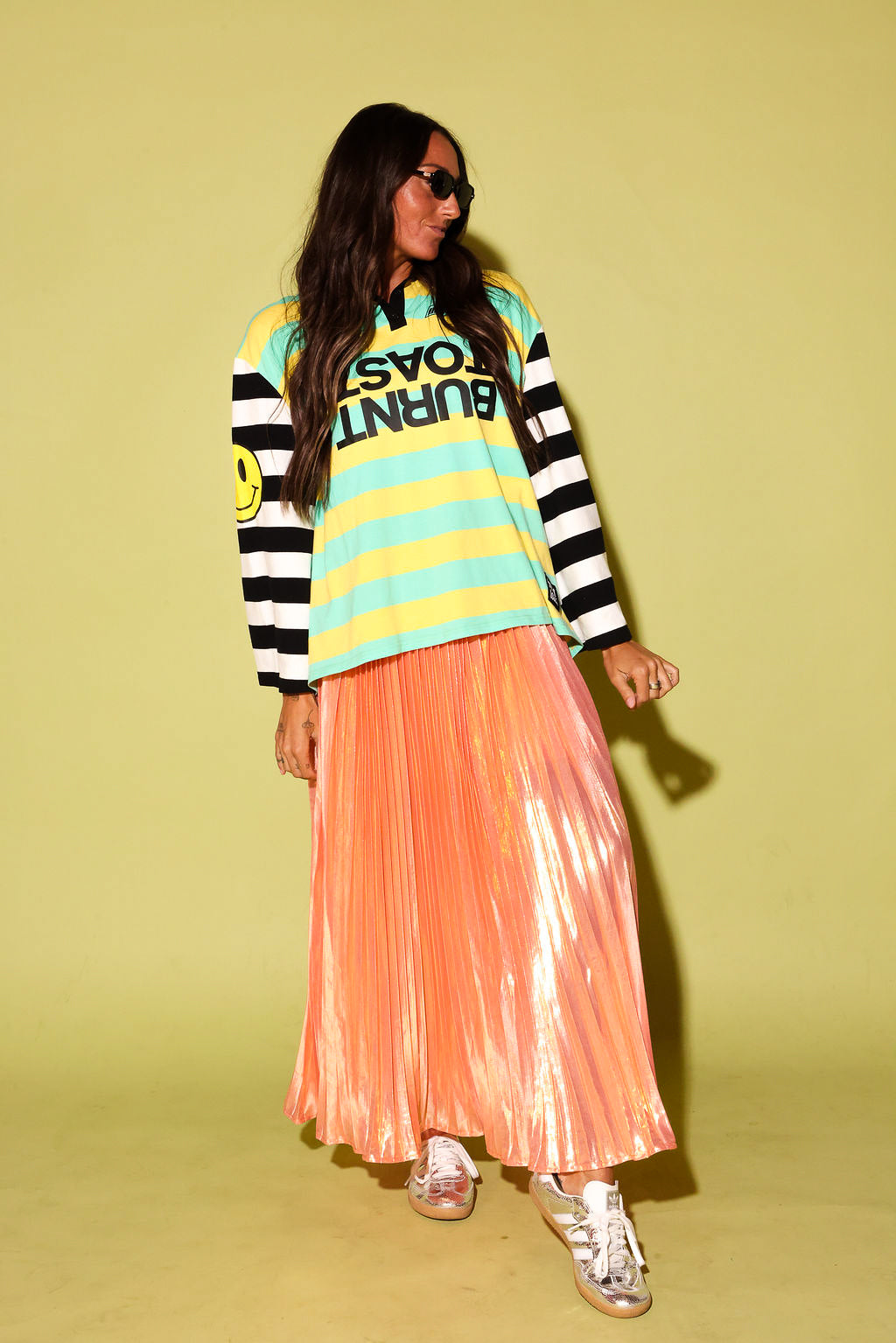 A model wearing a striped polo with a burnt toast logo and peach metallic plisse skirt, black beanie, and silver sneakers on a pale green background
