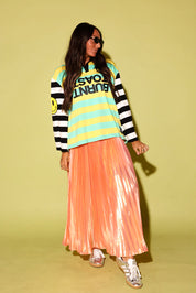 A model wearing a striped polo with a burnt toast logo and peach metallic plisse skirt, black beanie, and silver sneakers on a pale green background