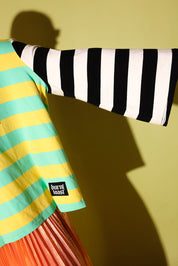 A close-up of a striped garment draped across a mannequin or form against a yellow-green wall. The visible fabric includes a black-and-white striped sleeve and a yellow-and-turquoise striped section with a small black brand patch reading 'burnt toast'.