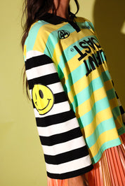 A close-up of a person wearing a colorful striped long-sleeve shirt with black and white horizontal stripes on one sleeve, yellow and mint green horizontal stripes on the body, a black collar, and a large yellow smiley face graphic on the sleeve.