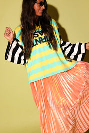 A model wearing a striped polo with a burnt toast logo and peach metallic plisse skirt, black beanie, on a pale green background