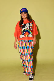 A woman modeling a vibrant orange graphic t-shirt with a cow and rainbow design, paired with colorful diamond-patterned pants, a blue baseball cap, and black sneakers, set against a solid pale yellow-green studio background.